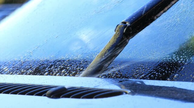 Close-up view of a car windshield being cleaned with a wiper, showcasing water droplets and dirt removal, camera pans across the scene highlighting the action flow