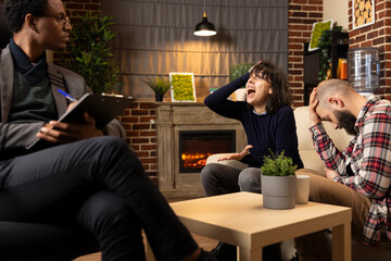 Tensed couple in therapy session facing conflict during marriage counseling. Relationship counselor mediates discussion as frustrated husband bows head and stressed wife holds hand to forehead.