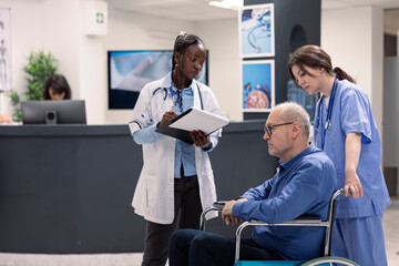 Obraz premium Caucasian nurse assists elderly wheelchair user during health appointment with african american doctor. Black female physician reviews medical notes and rehabilitation options with retired man.