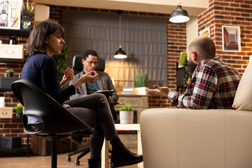 Fototapeta premium Anxious woman looks upset speaking to irritated husband during therapy session as black male psychiatrist writes down notes. Married couple struggling in counseling, arguing about emotional struggles.