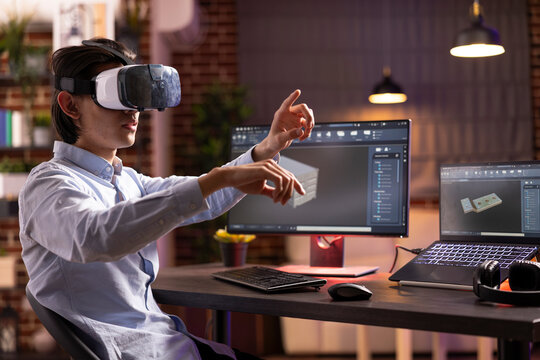 Male freelancer using virtual reality headset to visualize 3D product prototypes with CAD software on computer screens. Young asian man developing digital container design at home desk.