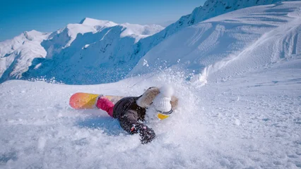 Fotobehang Extreme Sporten CLOSE UP: Snowboarder slides down the slope after a crash into soft snow while learning to carve. Rugged white mountain peaks fill the horizon, capturing both the challenge and fun of winter riding.  © helivideo