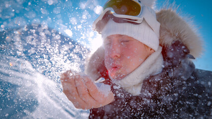 LENS FLARE, CLOSE UP, DOF, PORTRAIT: Playful woman blowing a handful of fresh snow toward the camera. Cloud of sparkling white crystals explodes into the sunlight, catching bright winter sunbeams.