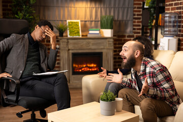 Psychologist holding clipboard with hand on forehead listens to couple during tense marriage counseling. Stressed counselor observes husband and wife sharing conflicts in brick wall therapy room.