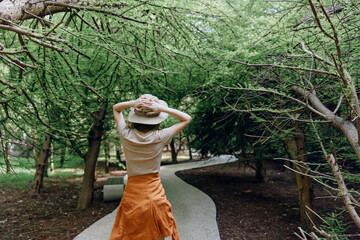 Woman in a hat walking along a winding path through a lush forest with tall trees, back view of a...