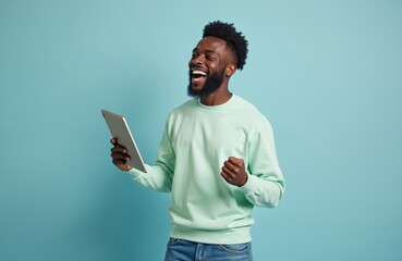 Happy african man holds tablet computer, laughs with joy, celebrates success online. Young guy with beard clenches fist, excited by good news, wins, achievement in studio.
