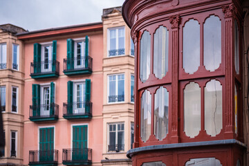 A close-up shot of a classic Bilbao apartment building facade, highlighting a distinctive reddish-brown rounded bay window contrasted with pale walls and green-shuttered balconies.