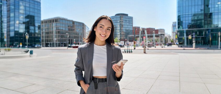 Young smiling elegant Asian busy business woman leader wearing suit standing in big city using cell phone platform applications. Smiling woman holding smartphone walking on street outdoors