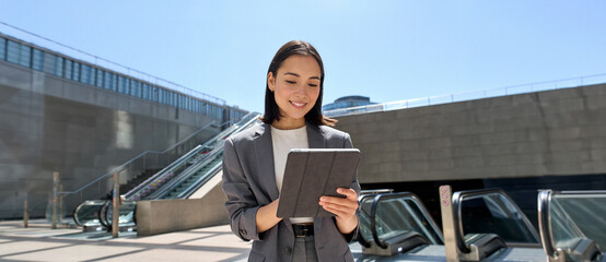 Young smiling Asian business woman entrepreneur wearing suit holding digital tablet standing in city metro using wireless internet technology on pad computer, working online in subway