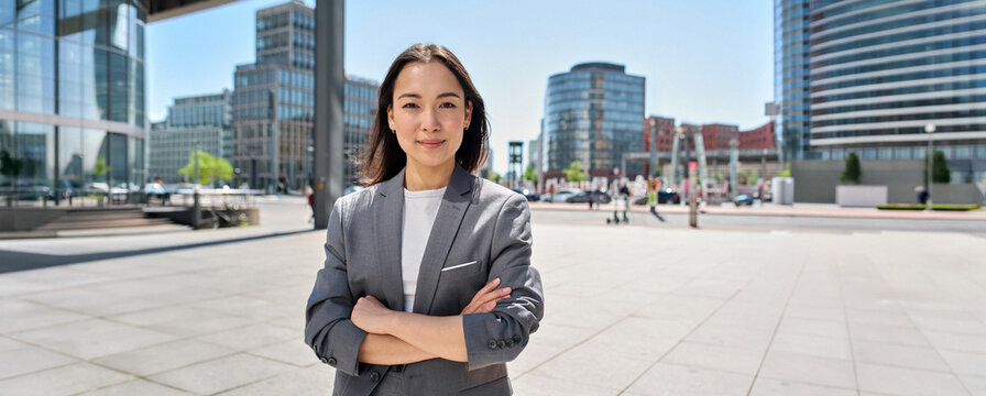 Young confident smiling Asian business woman standing on busy street, portrait. Proud successful female entrepreneur wearing suit posing with arms crossed looking at camera in big city outdoors