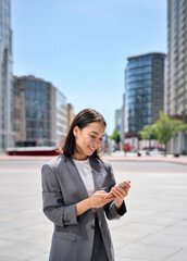 Young happy Asian business woman wearing suit holding mobile phone standing in big city on busy...