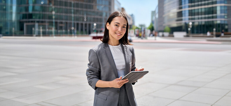 Young happy Asian business woman entrepreneur wearing suit holding digital tablet standing on big city busy street using smart business software tech for online work on pad computer outdoor, vertical - Powered by Adobe