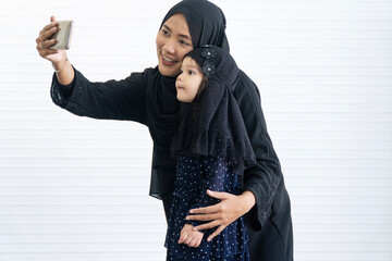 Happy Muslim mother hijab and daughter take selfie with smartphone white background. Loving portrait captures family bonds, modern communication, Islamic lifestyle technology, social media concept.