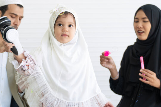 Cute Muslim toddler girl wearing white prayer hijab looking forward, standing between her parents in an indoor family setting. Represents happy family bonding and Islamic cultural education