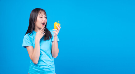 Surprised young Asian woman holding fresh yellow bell pepper with excitement on blue background. Beautiful healthy girl looking at organic vegetable with copy space for text.