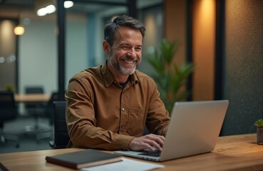 Fototapeta premium Smiling man with beard uses laptop in modern office after work hours. Busy professional types on computer at wooden desk. Focus on success and dedication.