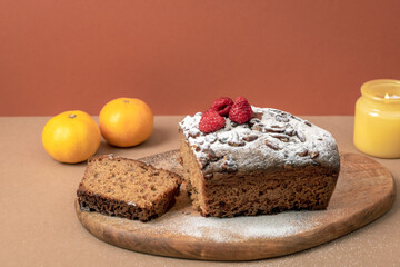Chocolate loaf cake with raspberries and sugar powder on brown table. Tangerines and yellow candle on the background. Closeup