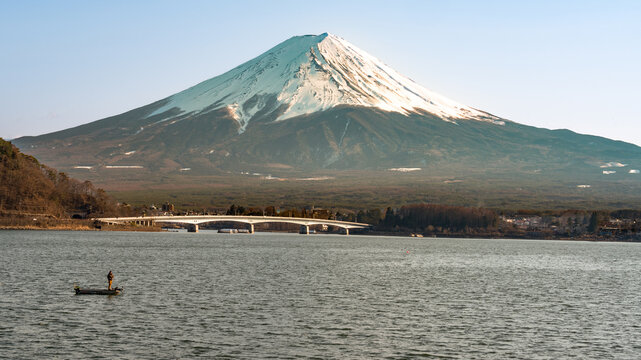 Mount Fuji overlooking Lake Kawaguchiko, Japan at sunrise with bridge and fishing boat with fisherman in the foreground.