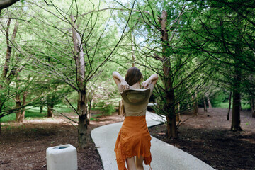 Woman walking on curved forest path between tall green trees, casual summer outfit and backpack,...