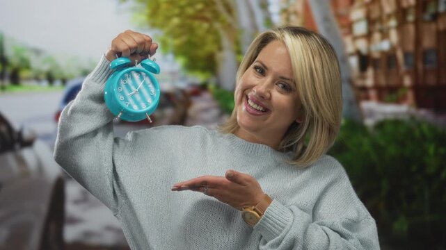 Woman holds turquoise alarm clock above head on urban street pointing at clock face while looking at camera; urgent reminder.