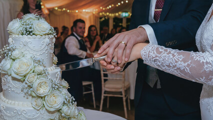 Bride and groom newlyweds holding hands and cutting their wedding cake at their reception. Friends and family in the background. Wedding rings. Floral cake design.