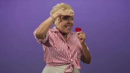 Elderly woman holding a red heart in one hand while shading her eyes with the other in a studio with purple backdrop; hopeful affection.