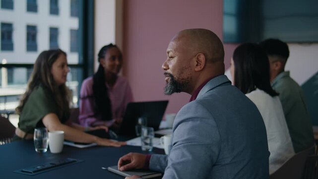 Smiling mature black businessman looking at camera while sitting with colleagues in boardroom at office