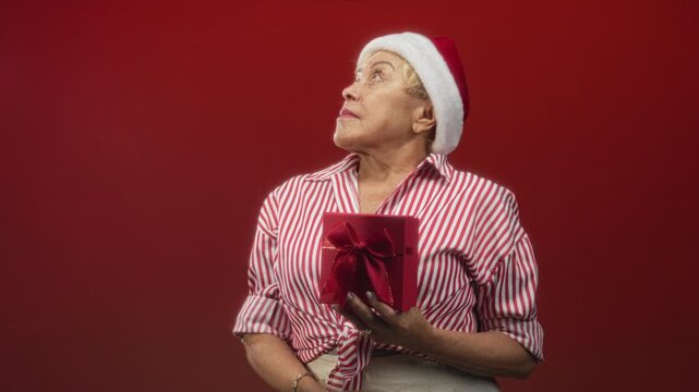 Woman wearing santa hat holding red gift box with hand on abdomen, looking up in studio against red backdrop; holiday nostalgia.