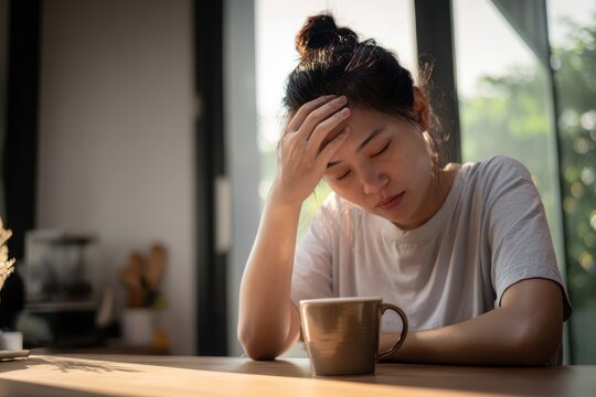 Stressed woman with a headache seeks relief with coffee at home, depicting burnout, mental health, and the need for self-care in today's demanding world