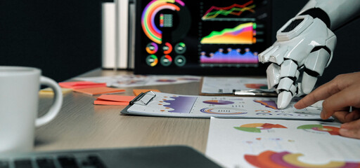 Futuristic robotic hand analyzing colorful presentation charts on a modern workspace table with electronic screens displaying data visualizations and graphs. Noogenesis