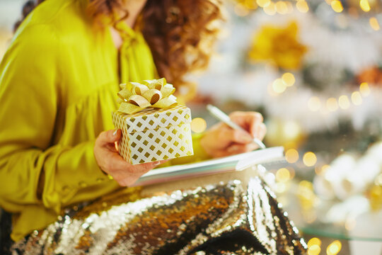 A woman with curly hair, wearing a shiny yellow blouse and sparkling pants, holds a gold-patterned gift box while writing in a notebook, set against a backdrop of warm bokeh lights.