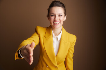 A confident businesswoman in a yellow blazer smiles warmly, extending her hand for a handshake, symbolizing a welcome, new partnership, or a successful business agreement.