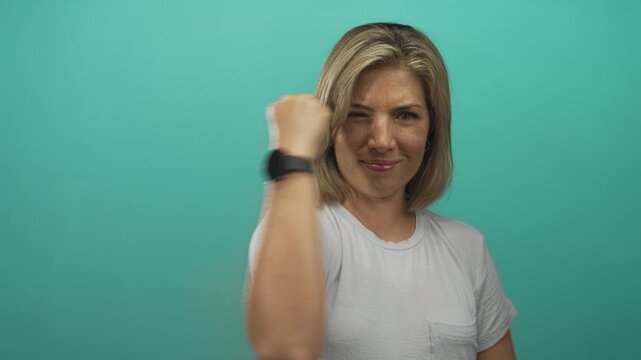 Young blonde woman displays victory sign with raised hand in turquoise studio; triumph confidence spirit.