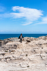 Woman walking a dog on a rocky cliffside with the blue ocean and sky in the background in Los Escullos beach in Spain