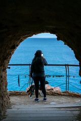 Woman with dog photographing Spanish coast view from cave at dawn