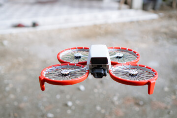 Small White Drone With Red Propeller Guards Hovers Outdoors In Daylight With Blurry Background