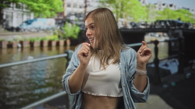 Young woman raises hand and smiles by canal on street during daytime while dancing; joy spontaneity.