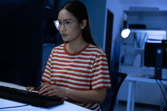 Asian young woman night work focused at computer wearing glasses and striped shirt in modern office with soft lamp glow