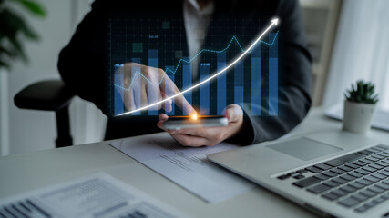A business professional engages with a smartphone, visualizing financial growth with digital graphs. The setting represents a modern office environment ideal for success. Impute