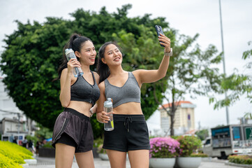 Asian woman friends in sportswear holding water and taking picture on phone after exercise in park.