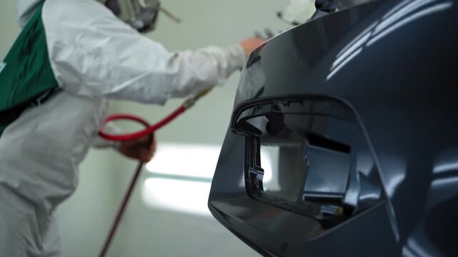 Auto technician spray gloss lacquer on car bumper inside clean, ventilated paint booth. Wearing protective coveralls and a respirator using a high precision spray gun.