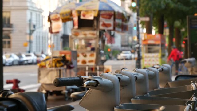 Manhattan Midtown street New York City, United States. Rental bicycle parking, Central Park West summer trees greenery. Bike for rent in row. People pedestrian on sidewalk pavement. Hot dog fast food.