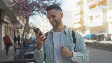 Young hispanic man celebrating outside on a sunny day, holding a phone and backpack with a joyful expression on an urban street filled with colorful trees and bustling activity.