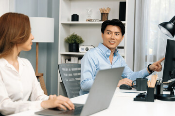 Smiling Asian man talking with woman colleague in planning marketing report project on laptop casual day while searching database on working desk at modern home office at side view image. Infobahn.