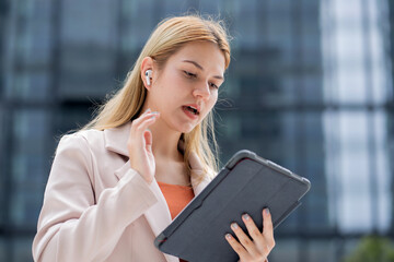 Professional young caucasian businesswoman talking using wireless earbud while holding digital tablet outdoors in city, business woman using technology for communication and success in urban.