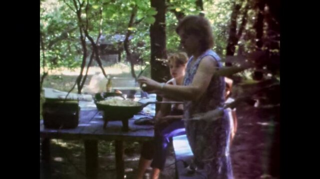 Woman cook food outdoors for family at summer wood picnic. Camp kitchen preparation, mess tent. Enjoying travel nature with children in green forest. Old film. Archival retro 1960s. Vintage archive