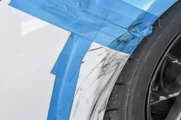 Close up of car damage with deep scratch on white fender. Blue masking tape protects vehicle panel during bodywork repair and paint job in professional garage