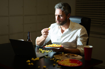 Man with laptop watching online video at night. Man working late eating junk food. Working late in office with snacks. Working late at night lifestyle. Night late concept for freelancers.