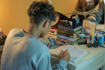 A young man studies at his desk, reading a book with full focus in a cozy indoor setting surrounded by stacks of books, landscape view