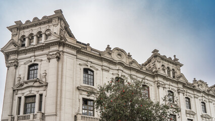 Fototapeta premium Old colonial Spanish architecture. A stone building with columns, arches, balustrades, balconies, and carvings on the facade. A green plant in the foreground. Peru. Historic Centre of Lima